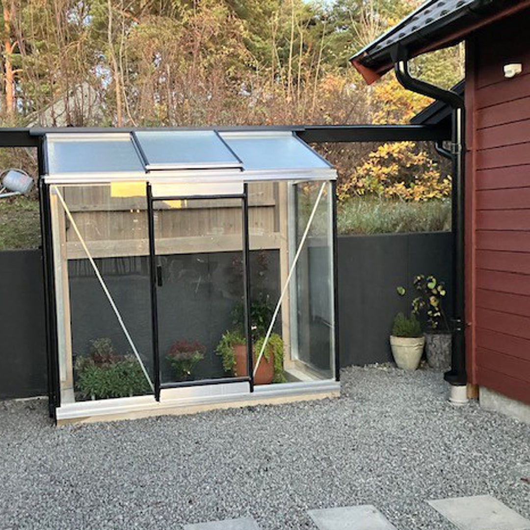 växthusbolaget växthus Miccolo 3 aluminium står mot ett plank  mellan 2 hus, grusgång